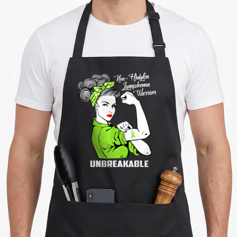 Non limes apron - Cartoon woman flexing arm with green ribbon and text Non-Hodgkin Lymphoma Warrior Unbreakable — shown on a black cotton canvas apron against a white background