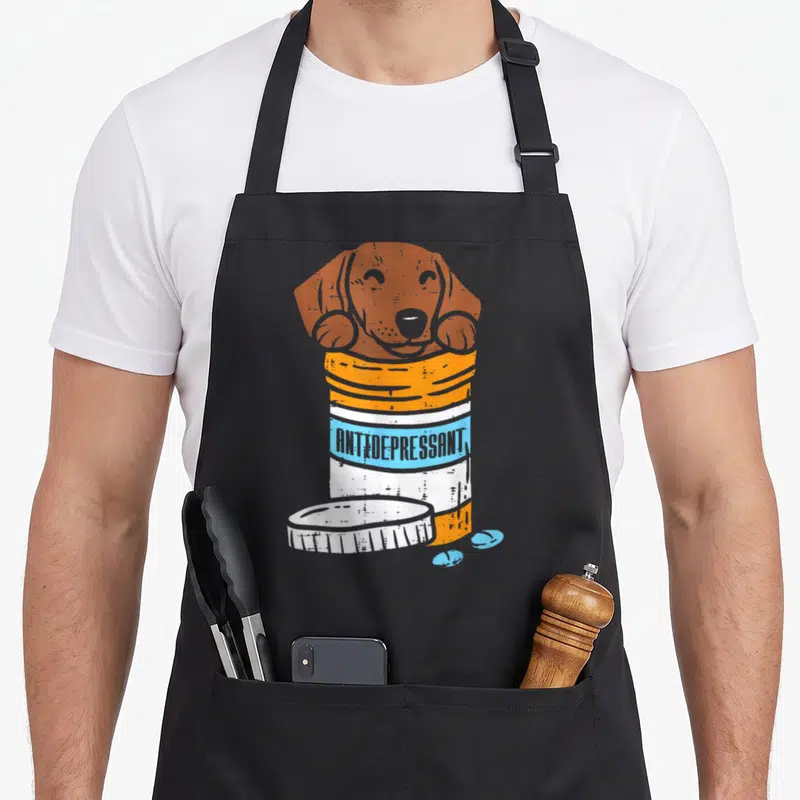Antidepressant sausage apron - Brown dachshund in orange pill bottle with blue pills and antidepressant text — shown on a black cotton canvas apron against a white background