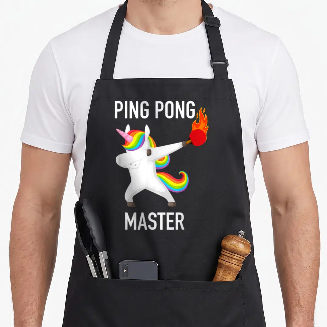 Ping master apron - Cartoon unicorn dabbing with flaming red paddle, PING PONG MASTER text above and below — shown on a black cotton canvas apron against a white background