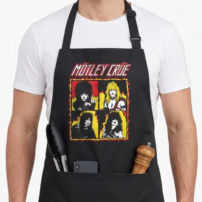 Devil e.a apron - Mötley Crüe text above four band portraits in a stylized flame frame with yellow and red accents — shown on a black cotton canvas apron against a white background