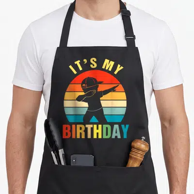 Cute dabbing birthday apron - Retro striped sun with grey dabbing silhouette and IT'S MY BIRTHDAY text — shown on a black cotton canvas apron against a white background