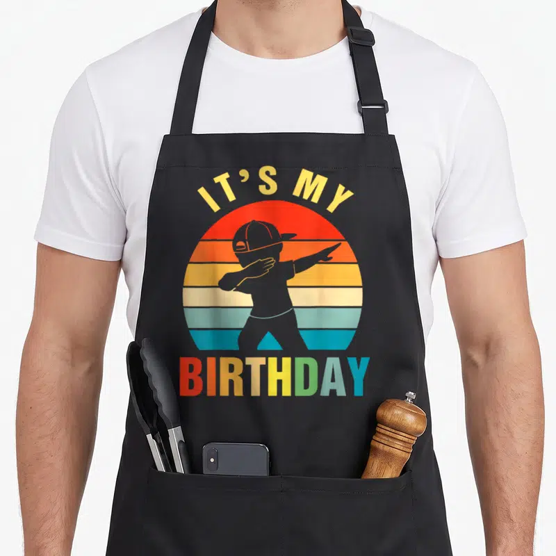 Cute dabbing birthday apron - Retro striped sun with grey dabbing silhouette and IT'S MY BIRTHDAY text — shown on a black cotton canvas apron against a white background