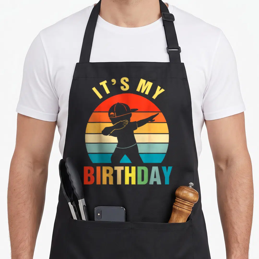Cute dabbing birthday apron - Retro striped sun with grey dabbing silhouette and IT'S MY BIRTHDAY text — shown on a black cotton canvas apron against a white background