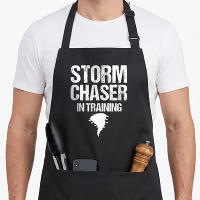 Storm training apron - White distressed text reading Storm Chaser In Training above a tornado silhouette — shown on a black cotton canvas apron against a white background
