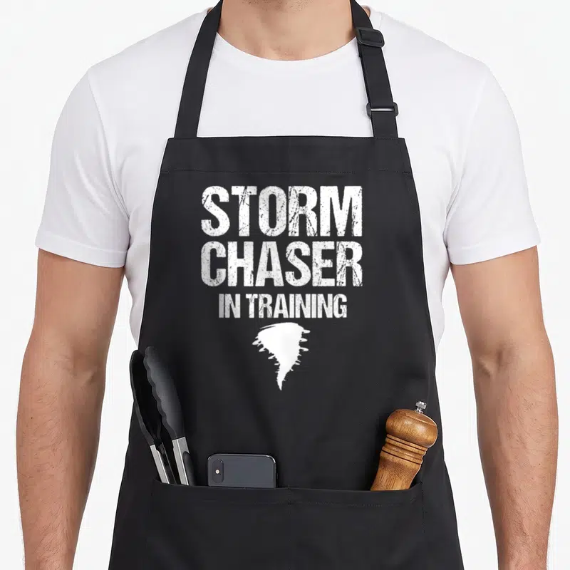 Storm training apron - White distressed text reading Storm Chaser In Training above a tornado silhouette — shown on a black cotton canvas apron against a white background