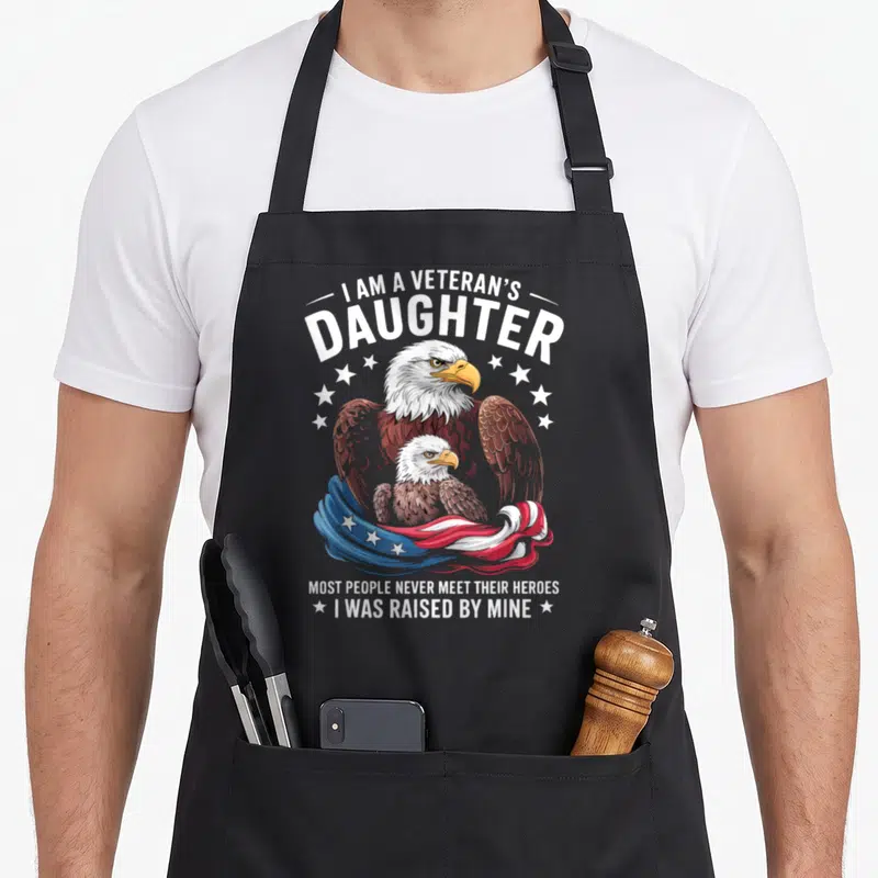 Veteran daughter people meet apron - Bald eagle and chick with patriotic flag and text about being raised by a veteran — shown on a black cotton canvas apron against a white background