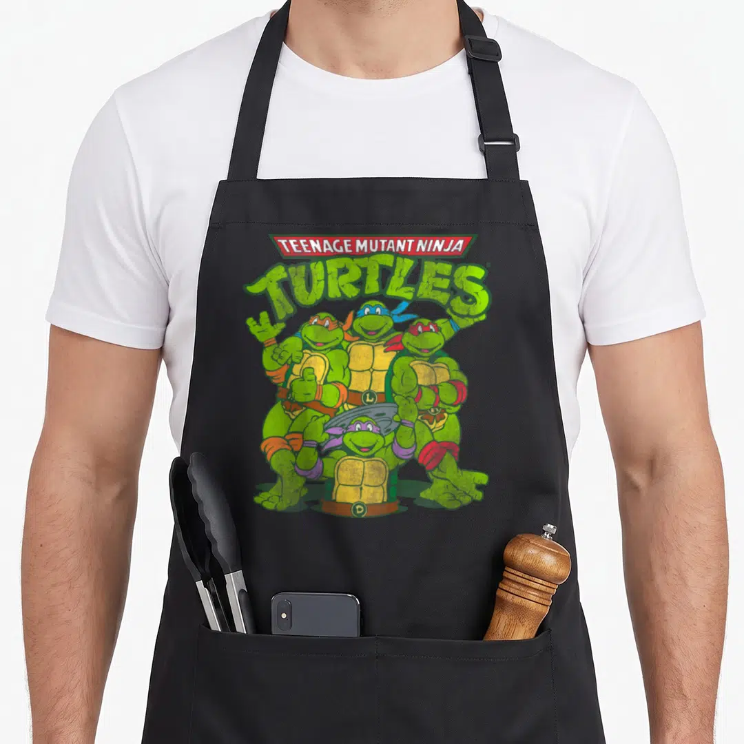 Distressed teenage cooking aprons - TEENAGE MUTANT NINJA TURTLES text above four turtles in green, red, blue, orange — shown on a black cotton canvas apron against a white background