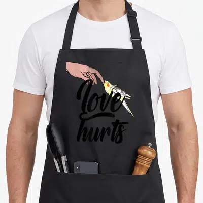 Funny cockatiel bird apron - Hand touching yellow grey cockatiel beak with Love Hurts black script text — shown on a black cotton canvas apron against a white background