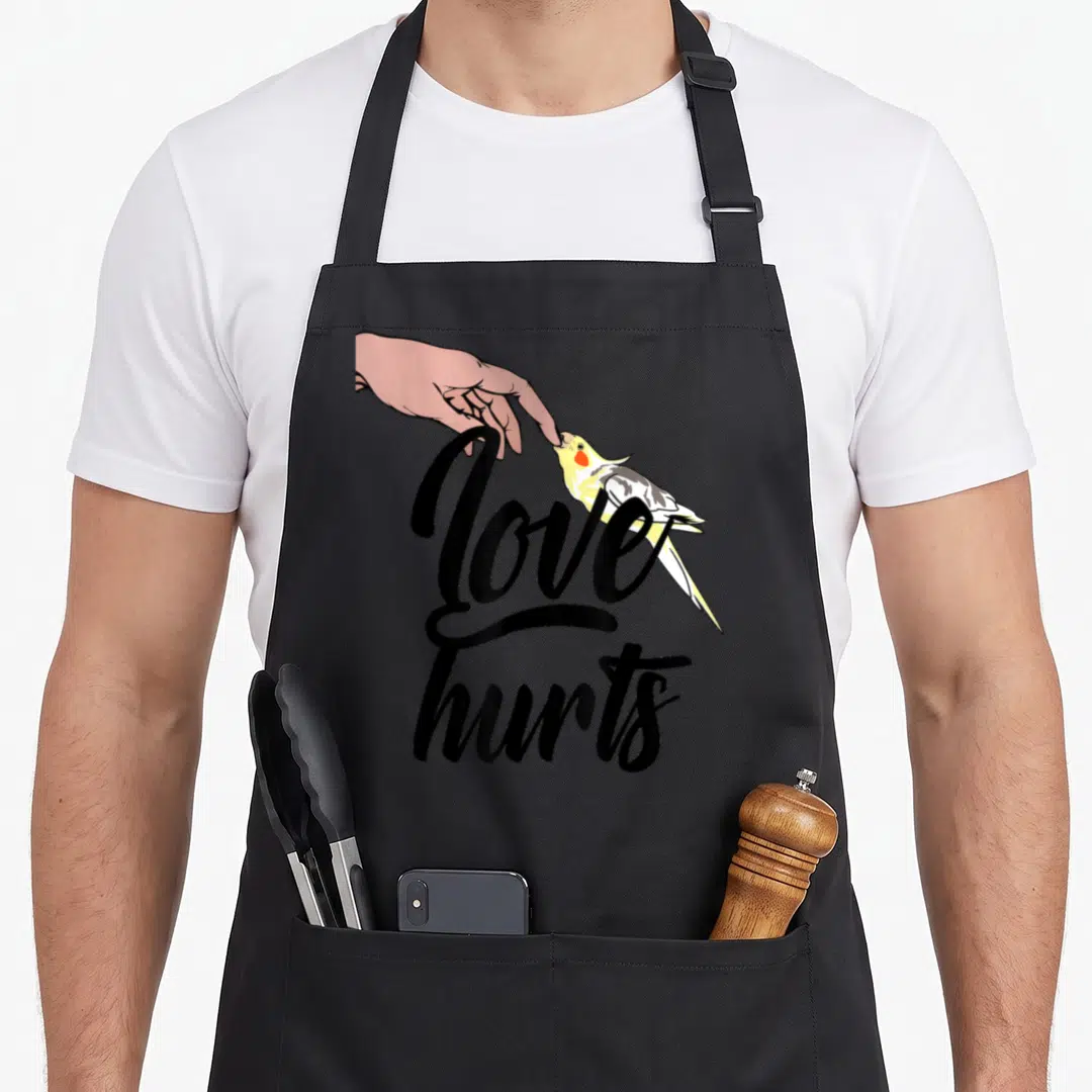 Funny cockatiel bird apron - Hand touching yellow grey cockatiel beak with Love Hurts black script text — shown on a black cotton canvas apron against a white background