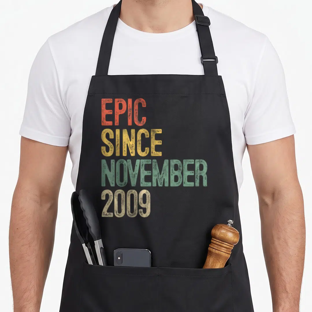 Epic november birthday apron - Stacked distressed text EPIC SINCE NOVEMBER 2009 in orange yellow green beige — shown on a black cotton canvas apron against a white background
