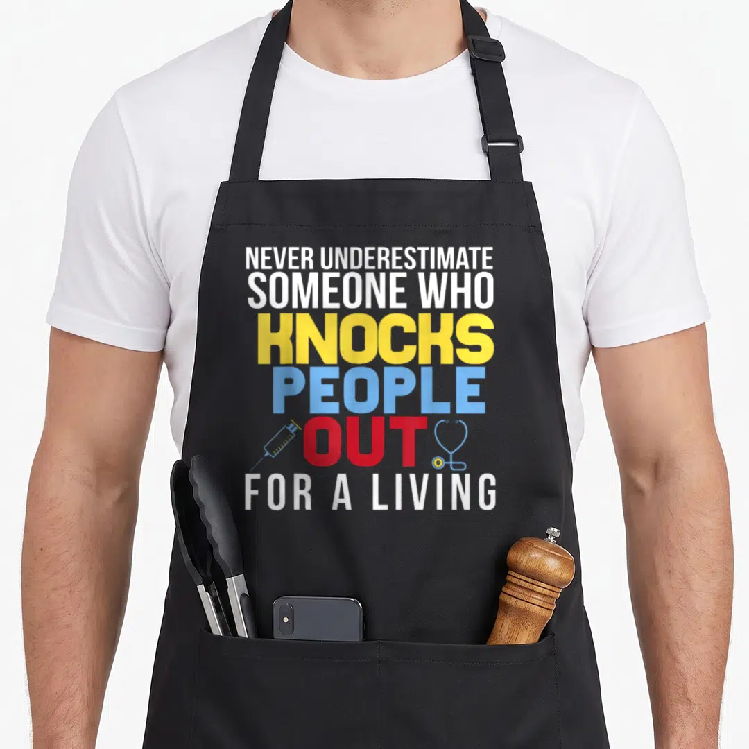 Crna nurse apron - Stacked text NEVER UNDERESTIMATE KNOCKS PEOPLE OUT FOR A LIVING with syringe and stethoscope — shown on a black cotton canvas apron against a white background