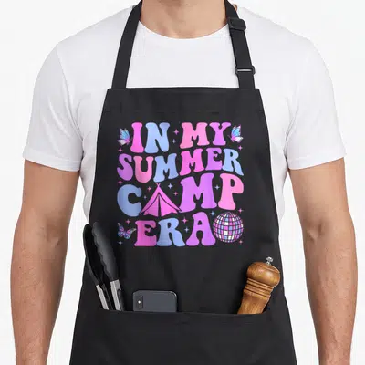 Groovy camp chef apron - Bubbly pink blue purple text IN MY SUMMER CAMP ERA with tent disco ball butterflies — shown on a black cotton canvas apron against a white background