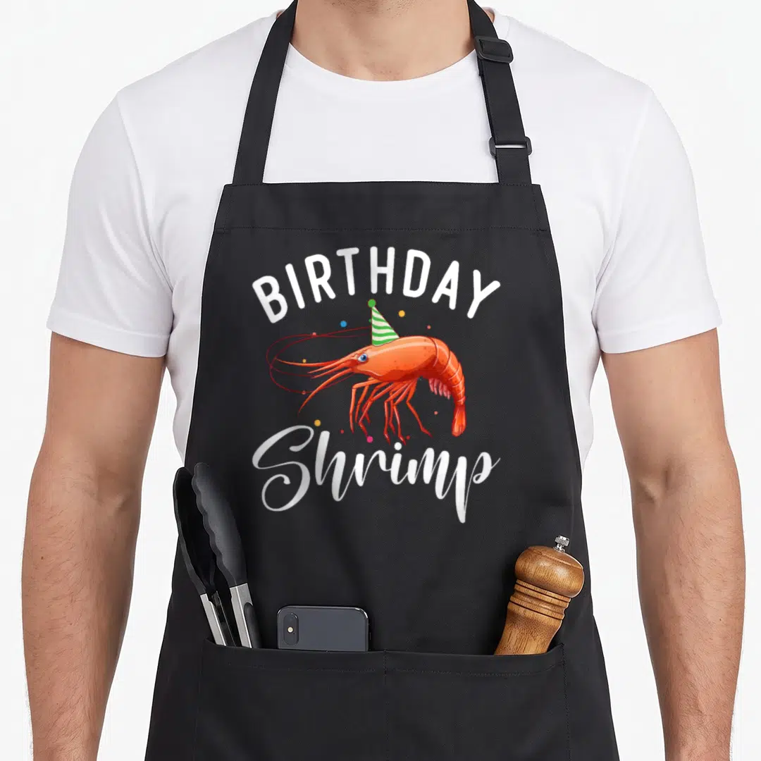 Shrimp birthday apron - Cartoon orange shrimp wearing green striped hat with BIRTHDAY Shrimp text and confetti — shown on a black cotton canvas apron against a white background