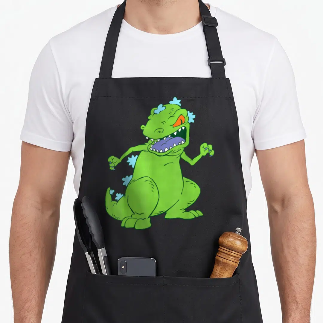 Reptar novelty apron - Green cartoon monster with blue spikes, purple tongue, orange eyes, roaring pose — shown on a black cotton canvas apron against a white background