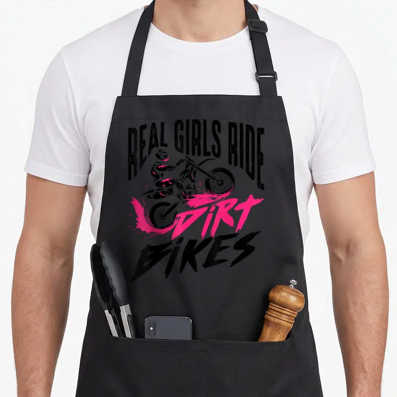 Racer bike apron - Real Girls Ride Dirt Bikes text with rider and pink splash — shown on a black cotton canvas apron against a white background