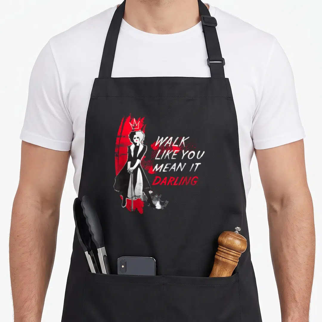 Cruella cruller apron - Woman in black dress with red splatter background and text Walk Like You Mean It Darling — shown on a black cotton canvas apron against a white background