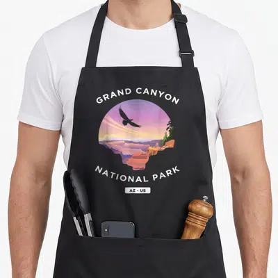 Grand us aprons - Retro Grand Canyon circular landscape with purple sky, eagle silhouette, and white arched text — shown on a black cotton canvas apron against a white background
