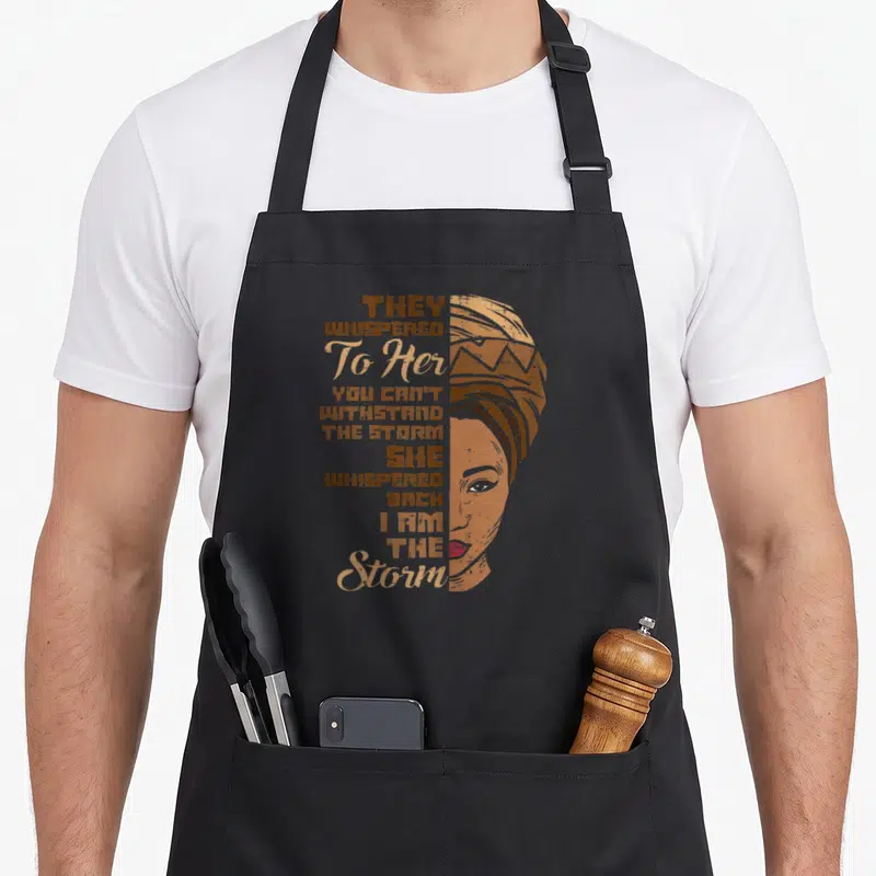 Storm queen apron - Distressed illustration of woman in headwrap with vertical quote They Whispered To Her You Can't — shown on a black cotton canvas apron against a white background