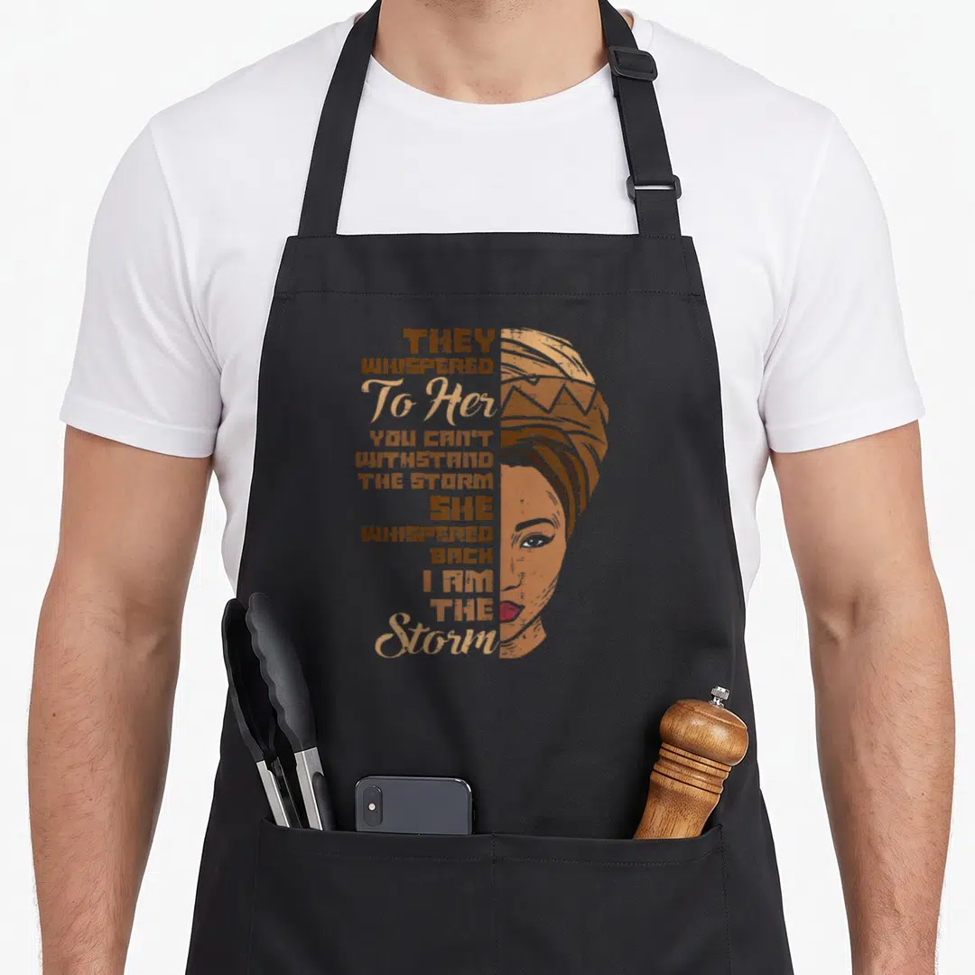 Storm queen apron - Distressed illustration of woman in headwrap with vertical quote They Whispered To Her You Can't — shown on a black cotton canvas apron against a white background