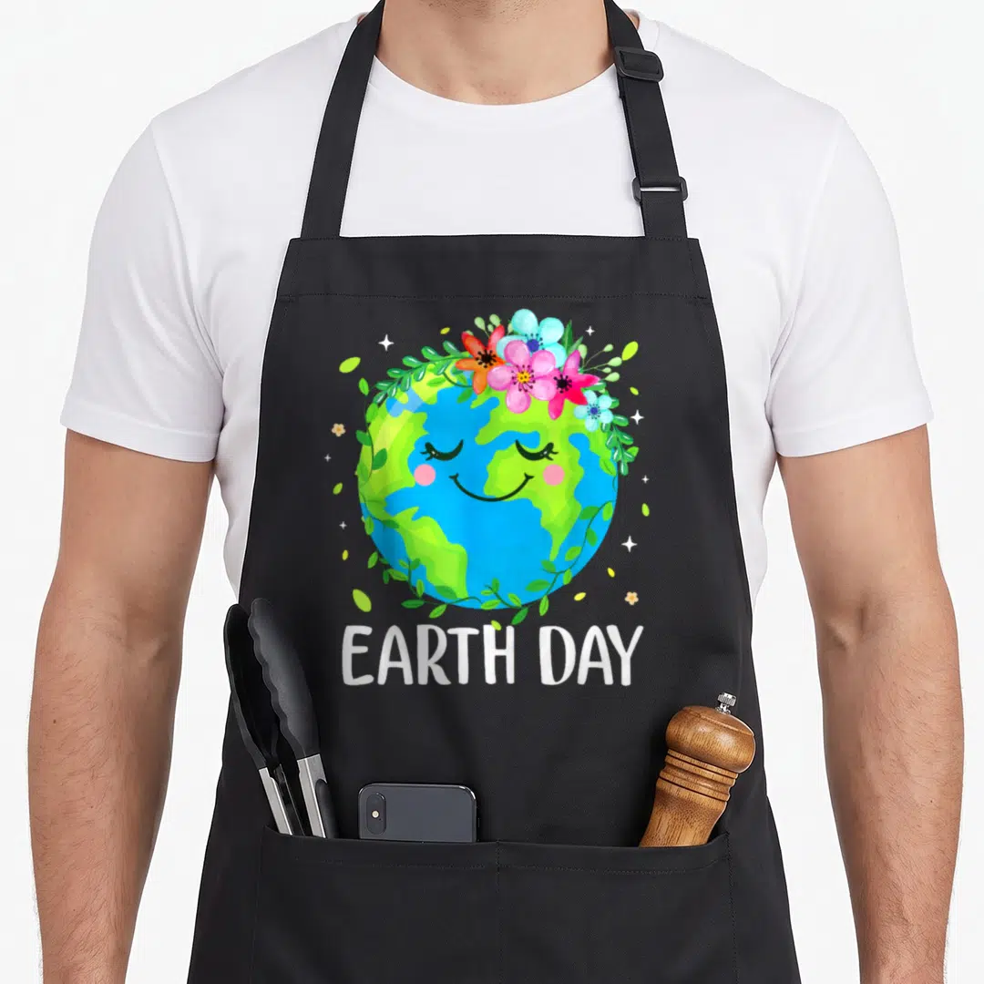 Earth floral apron - Smiling blue green Earth with pink flower crown and white EARTH DAY text below — shown on a black cotton canvas apron against a white background