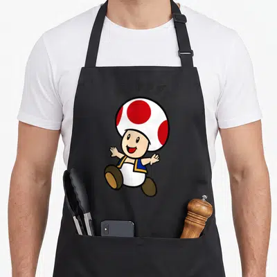 Stay toad apron - Cartoon Toad character with arched HAPPY! text in white red blue and dark grey — shown on a black cotton canvas apron against a white background