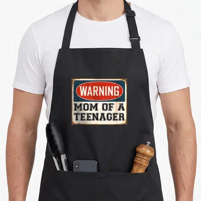 Warning mom apron - Rusty rectangular sign with red oval WARNING text and black MOM OF A TEENAGER lettering on white — shown on a black cotton canvas apron against a white background