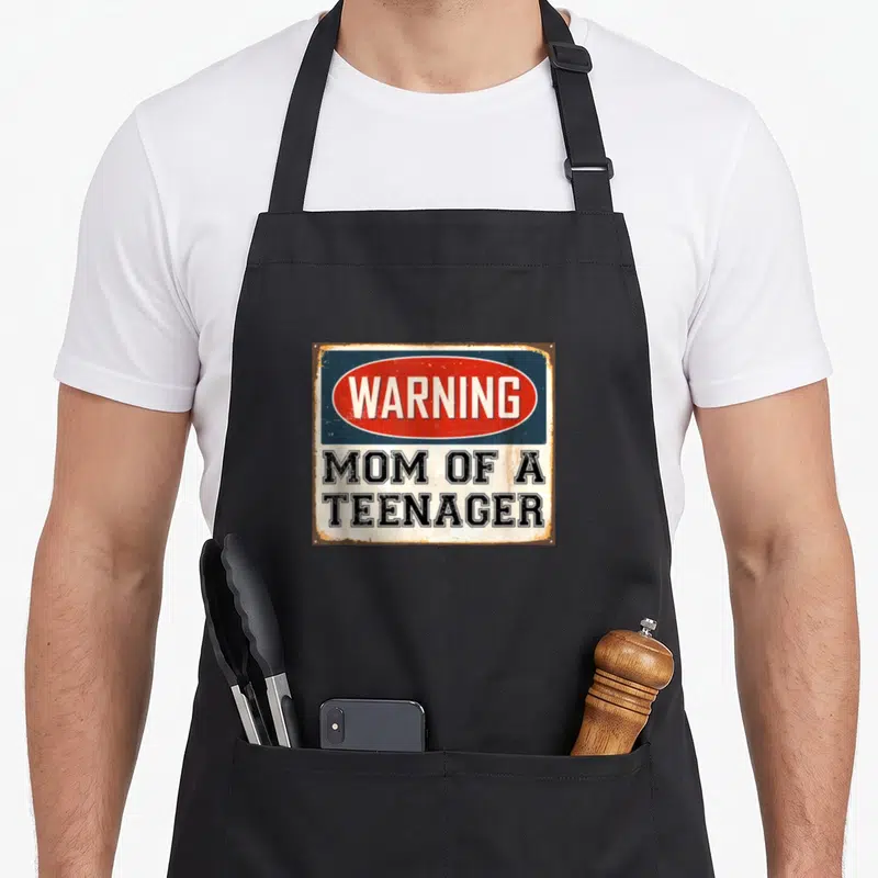 Warning mom apron - Rusty rectangular sign with red oval WARNING text and black MOM OF A TEENAGER lettering on white — shown on a black cotton canvas apron against a white background