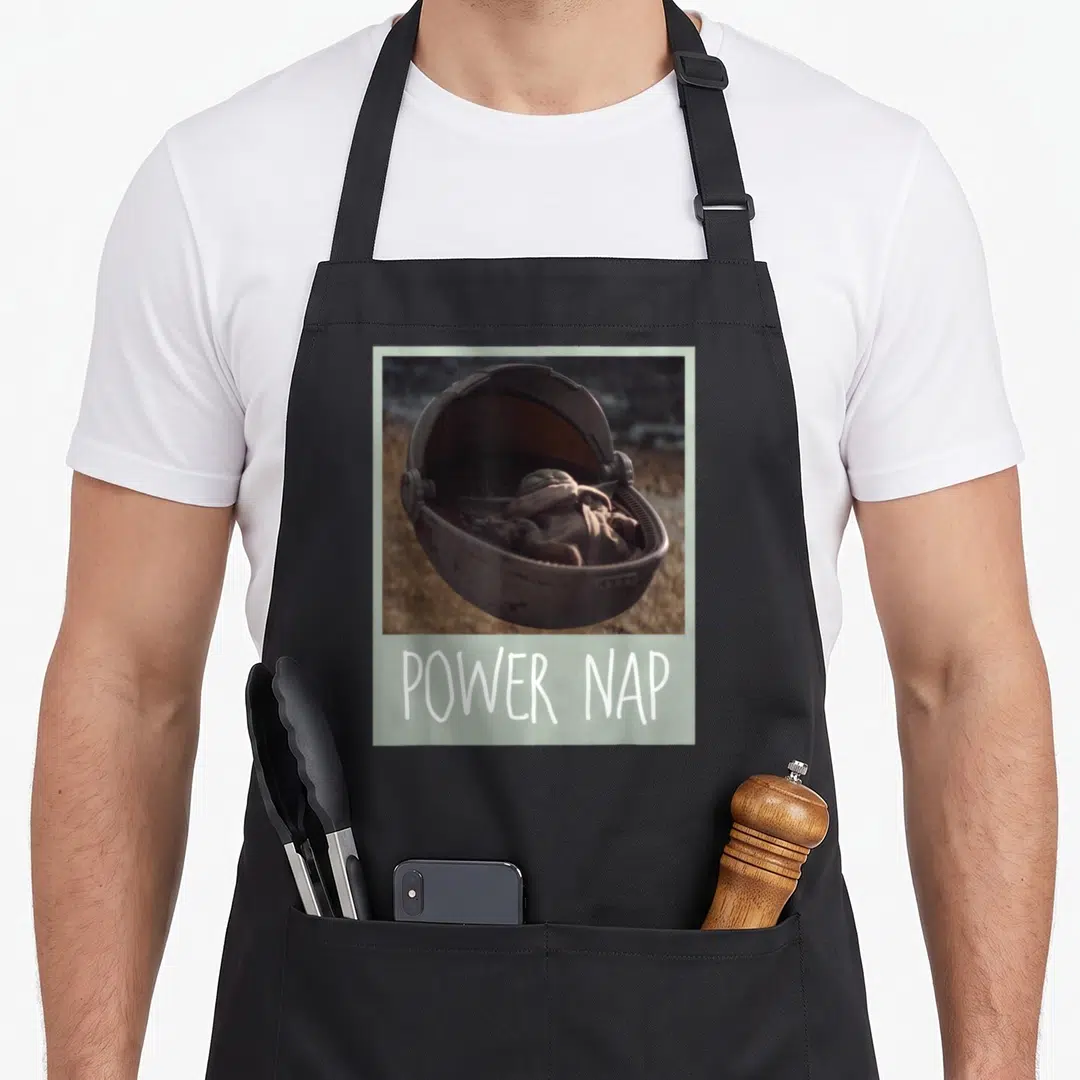 Power meme apron - Character sleeping in a floating pod above white POWER NAP text inside a light green border — shown on a black cotton canvas apron against a white background