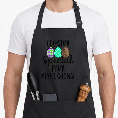 Eggstra teacher apron - Stacked text EGGSTRA Special PARA PROFESSIONAL above three patterned eggs in plaid, lattice — shown on a black cotton canvas apron against a white background
