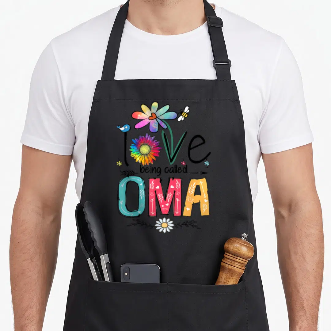 Called daisy apron - Rainbow flower and bird graphic with text love being called OMA on black fabric — shown on a black cotton canvas apron against a white background