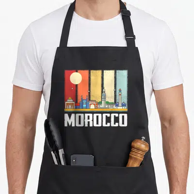Morocco african apron - Retro multicolored striped skyline with sun, buildings, lighthouse, and white MOROCCO text — shown on a black cotton canvas apron against a white background