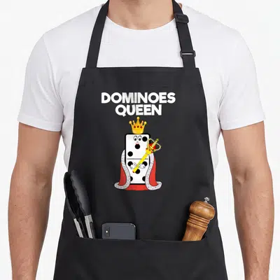 Dominoe queen apron - Cartoon domino character wearing a gold crown and red cape holding a scepter under text — shown on a black cotton canvas apron against a white background