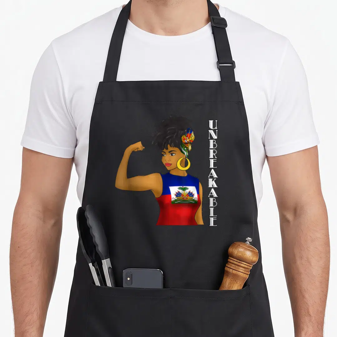Proud haitian apron - Cartoon woman flexing arm with vertical UNBREAKABLE text in blue red gold brown — shown on a black cotton canvas apron against a white background