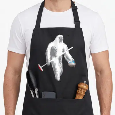 Curling bigfoot apron - White Yeti holding curling broom with red head and blue handled stone — shown on a black cotton canvas apron against a white background