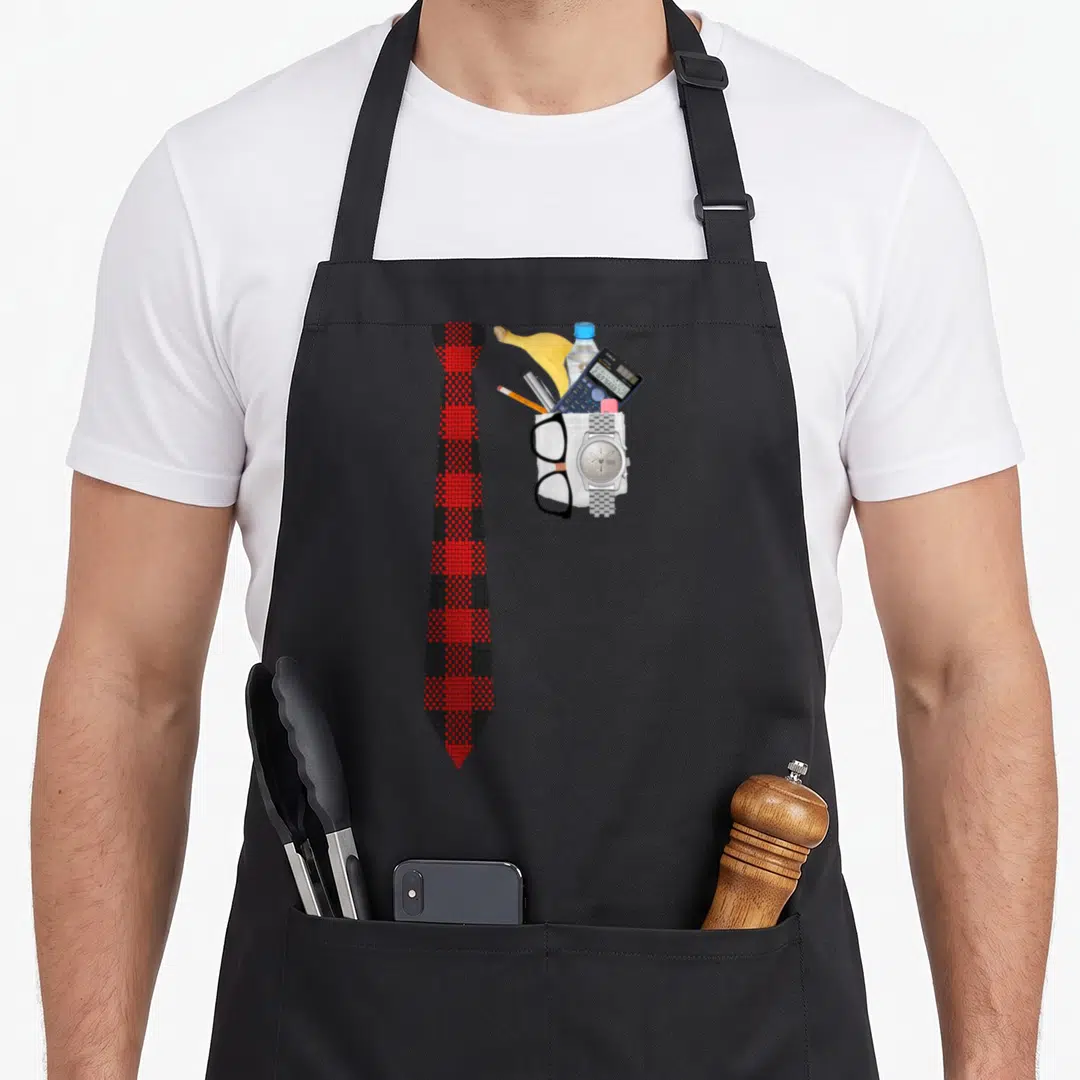 Halloween geek aprons - Red black plaid tie beside white bag with calculator banana and glasses — shown on a black cotton canvas apron against a white background
