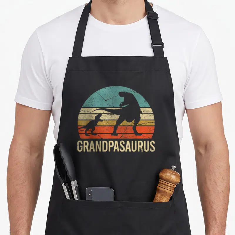 Dinosaur father day apron - Retro teal and orange sunset with dinosaur silhouettes and Grandpasaurus text — shown on a black cotton canvas apron against a white background