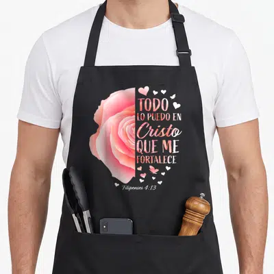 Christian spanish bible verse apron - Pink rose graphic next to stacked Spanish text and small hearts on grey background — shown on a black cotton canvas apron against a white background