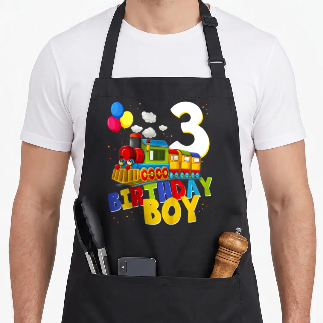 Train birthday apron - Cartoon train with balloons, large number 3, and BIRTHDAY BOY text in red blue yellow green white — shown on a black cotton canvas apron against a white background
