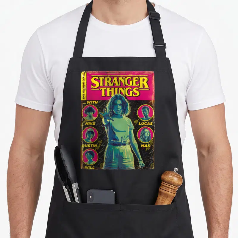 Shot stranger apron - Retro Stranger Things comic cover with girl and four named character portraits in magenta — shown on a black cotton canvas apron against a white background