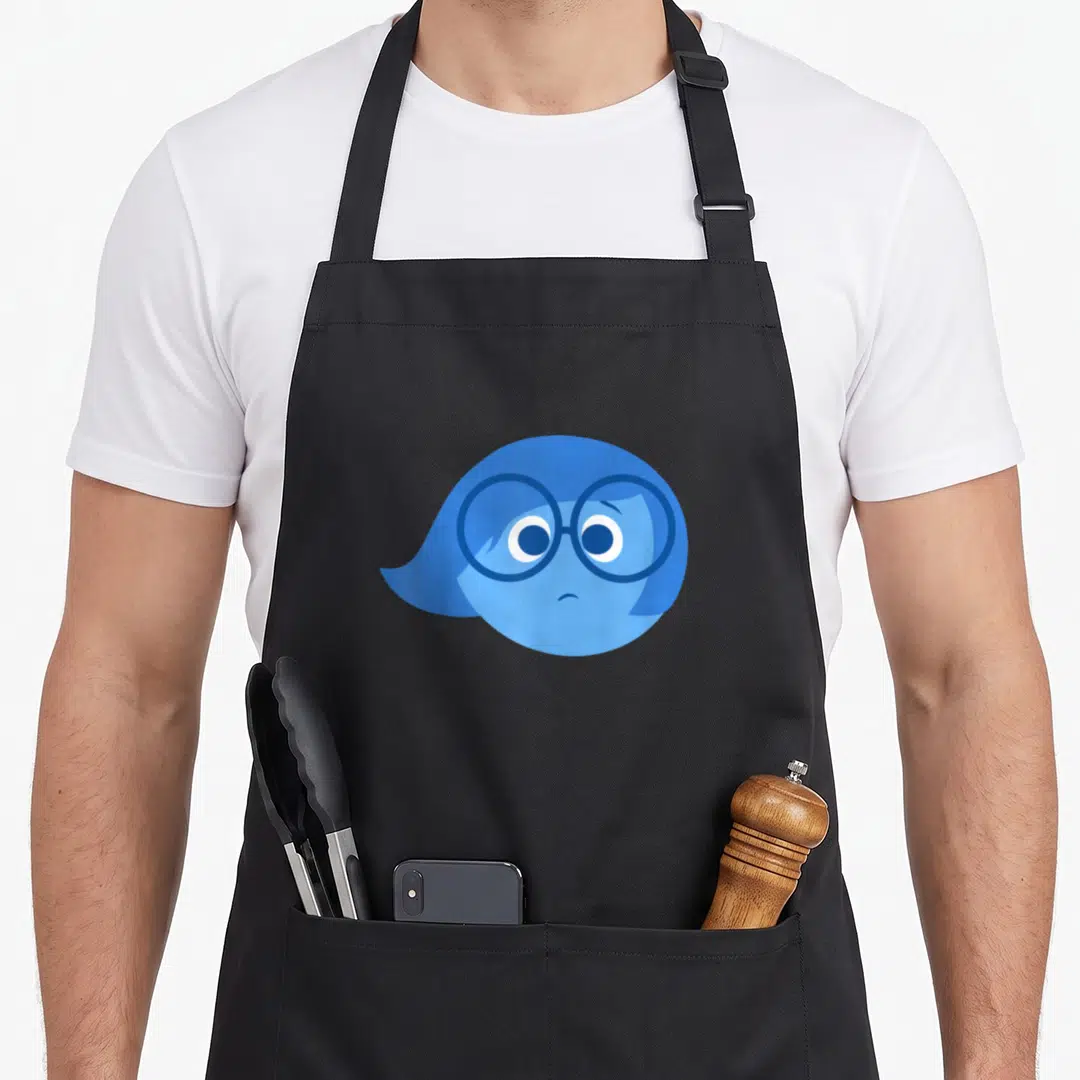 Sadness novelty apron - Blue cartoon character head with round glasses and frown on black background — shown on a black cotton canvas apron against a white background