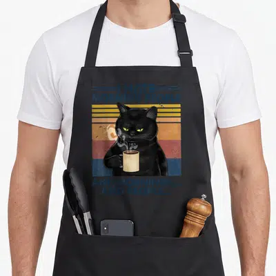 Grumpy coffee aprons - Black cat holding steaming mug with I Hate Morning People text on retro stripes — shown on a black cotton canvas apron against a white background