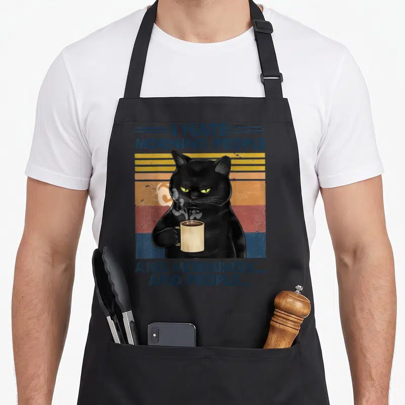 Grumpy coffee aprons - Black cat holding steaming mug with I Hate Morning People text on retro stripes — shown on a black cotton canvas apron against a white background