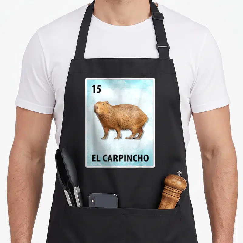 Carpincho mexican apron - Realistic brown capybara on light blue card with 15 and EL CARPINCHO text — shown on a black cotton canvas apron against a white background