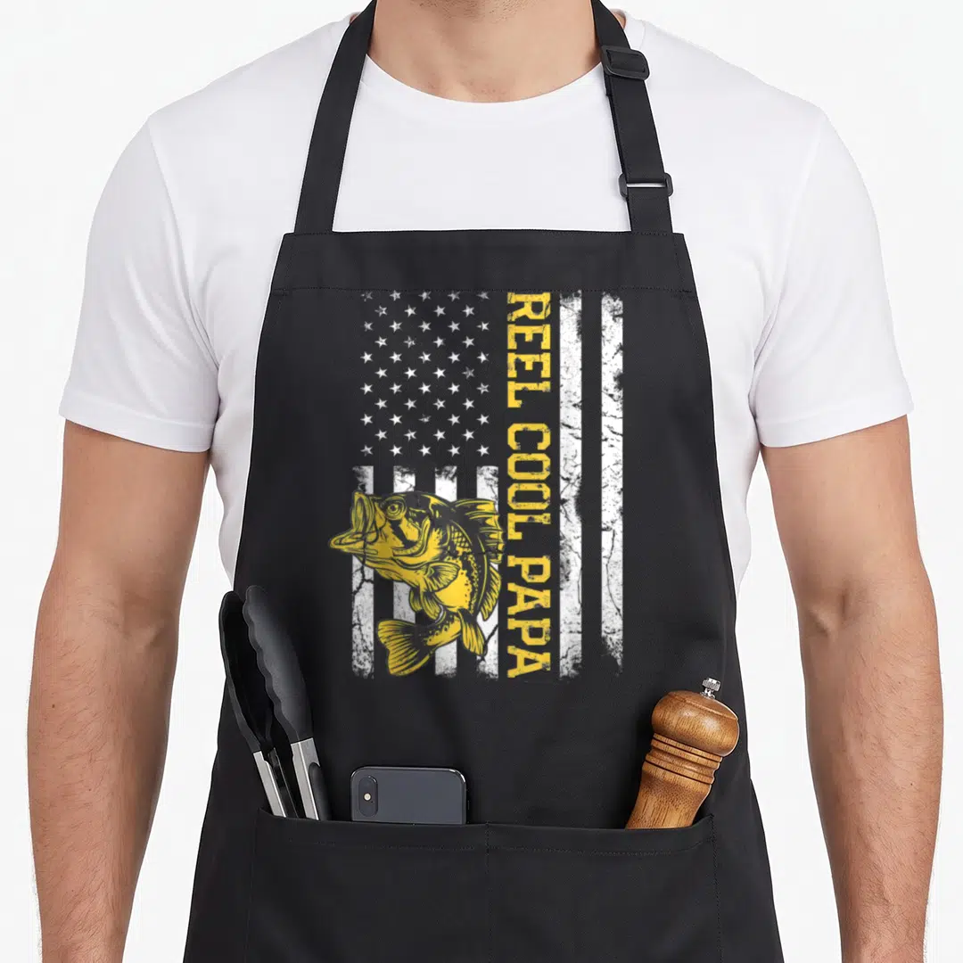 Reel dad joke apron - Yellow fish and vertical REEL COOL PAPA text over white stripes and stars — shown on a black cotton canvas apron against a white background