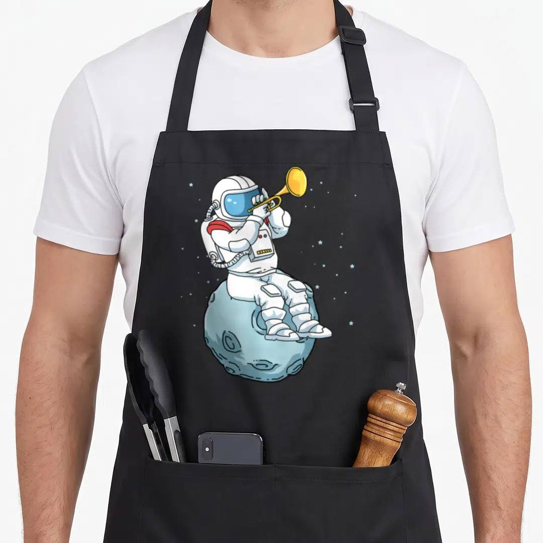 Funny player trumpet apron - Cartoon astronaut in white suit playing yellow trumpet while sitting on a light blue moon — shown on a black cotton canvas apron against a white background