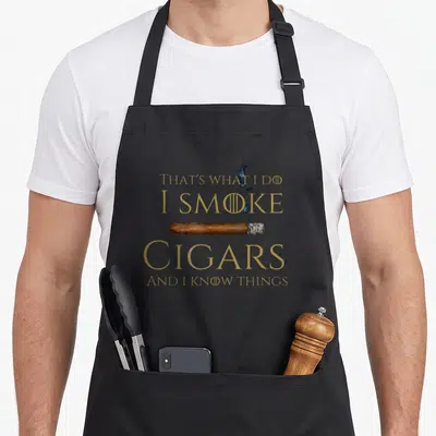 Cigar smoke smoker apron - Gold text reading I SMOKE CIGARS AND I KNOW THINGS surrounding a brown cigar with blue smoke — shown on a black cotton canvas apron against a white background
