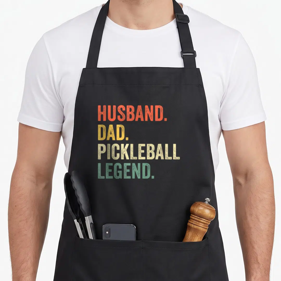 Pickleball husband apron - Stacked text HUSBAND. DAD. PICKLEBALL LEGEND. in orange yellow beige and green distressed — shown on a black cotton canvas apron against a white background