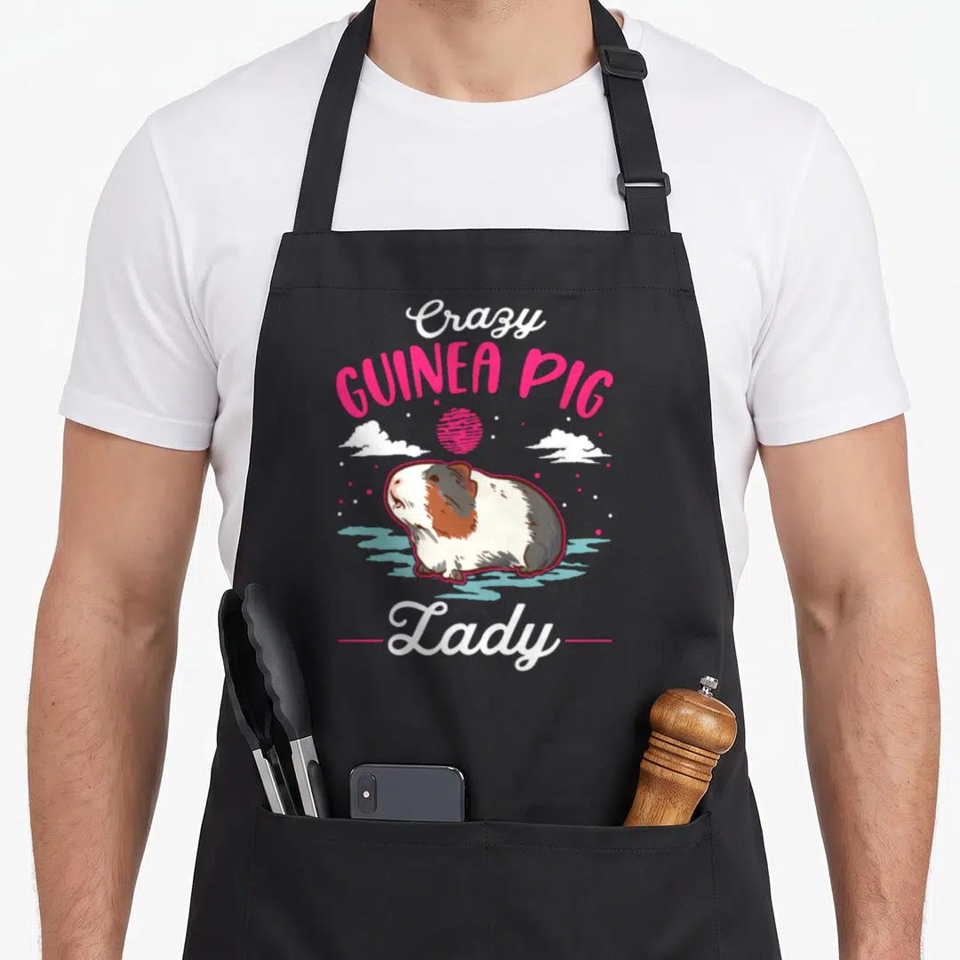 Cute pig animal crazy guinea lady apron - Cartoon guinea pig on blue water with pink sun and stacked text reading Crazy — shown on a black cotton canvas apron against a white background