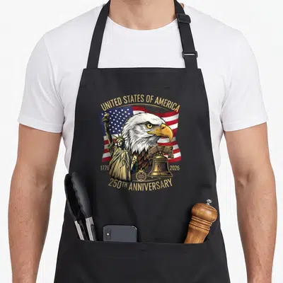 Birthday eagles apron - Bald eagle head over USA flag with Statue of Liberty and Liberty Bell in gold and patriotic — shown on a black cotton canvas apron against a white background