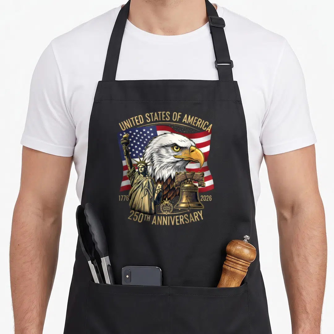 Birthday eagles apron - Bald eagle head over USA flag with Statue of Liberty and Liberty Bell in gold and patriotic — shown on a black cotton canvas apron against a white background
